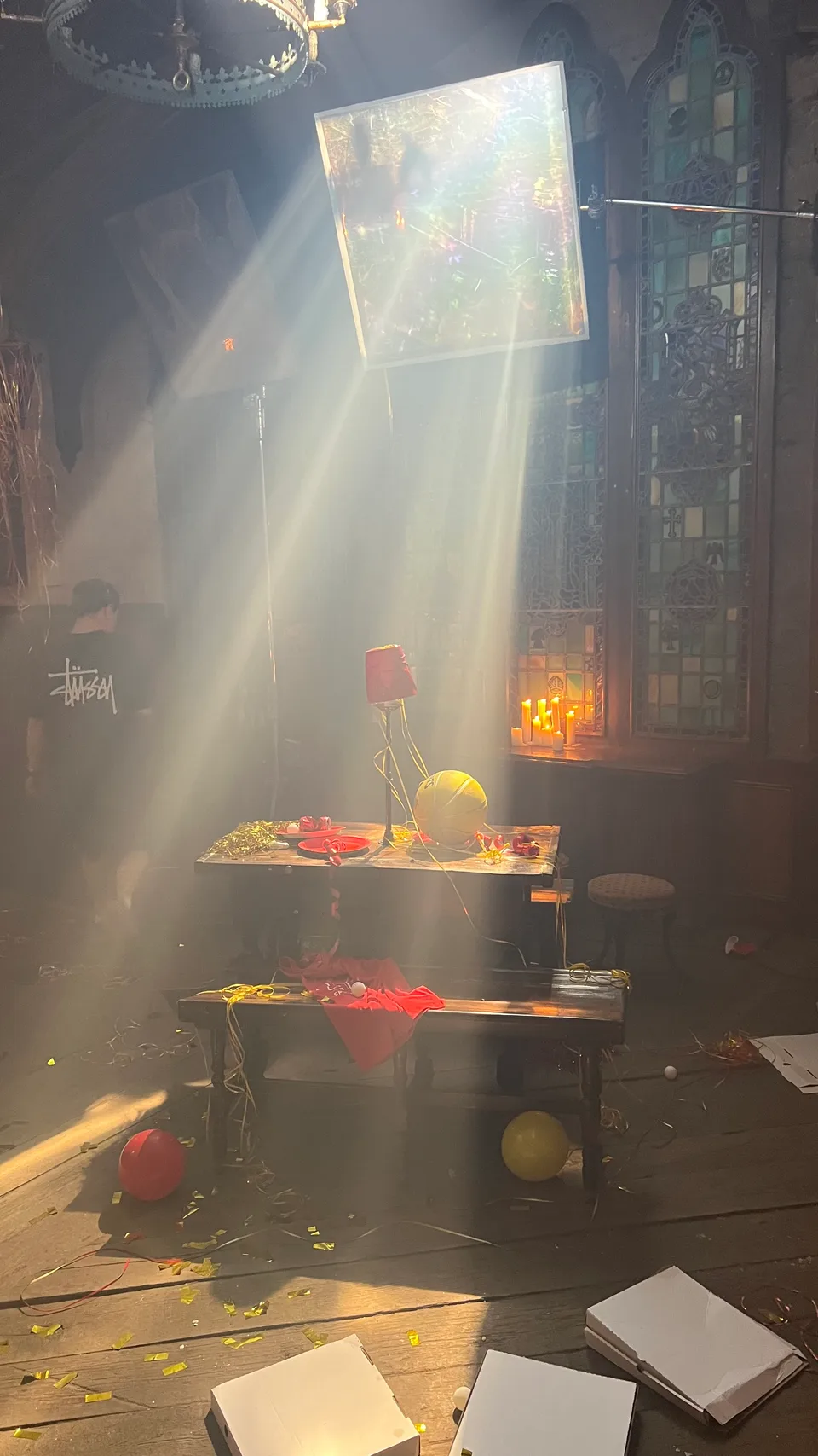 Behind the scenes: a dressed table, candles and props beneath shafts of light, with film equipment visible.