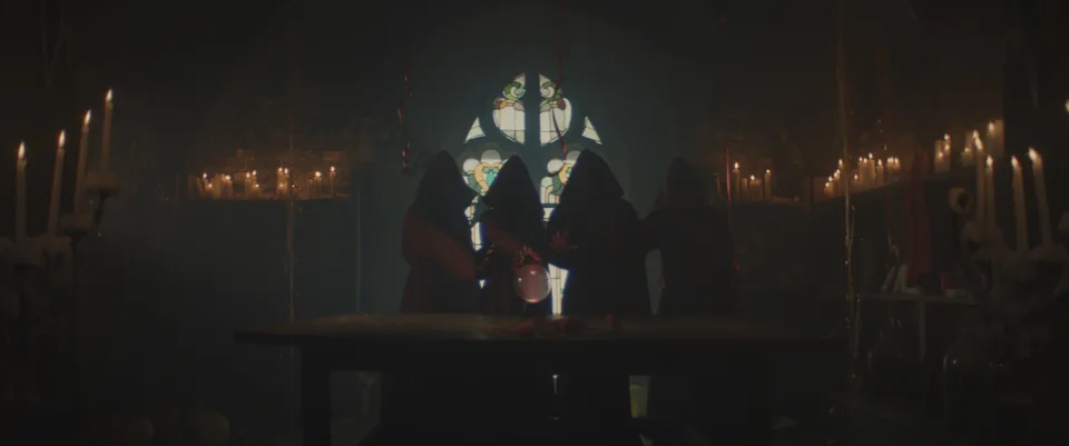 Two figures face each other across a table in a candlelit room with a pointed window.