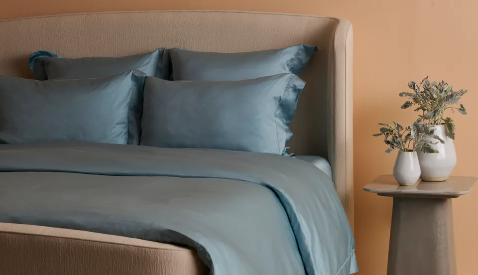 A bedroom set with blue bedding, a padded headboard and flowers on a bedside table.