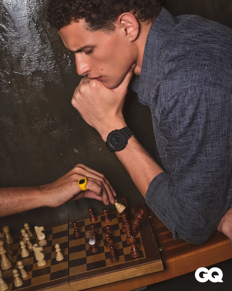 A model wearing a watch leans over a chessboard on a dark table.