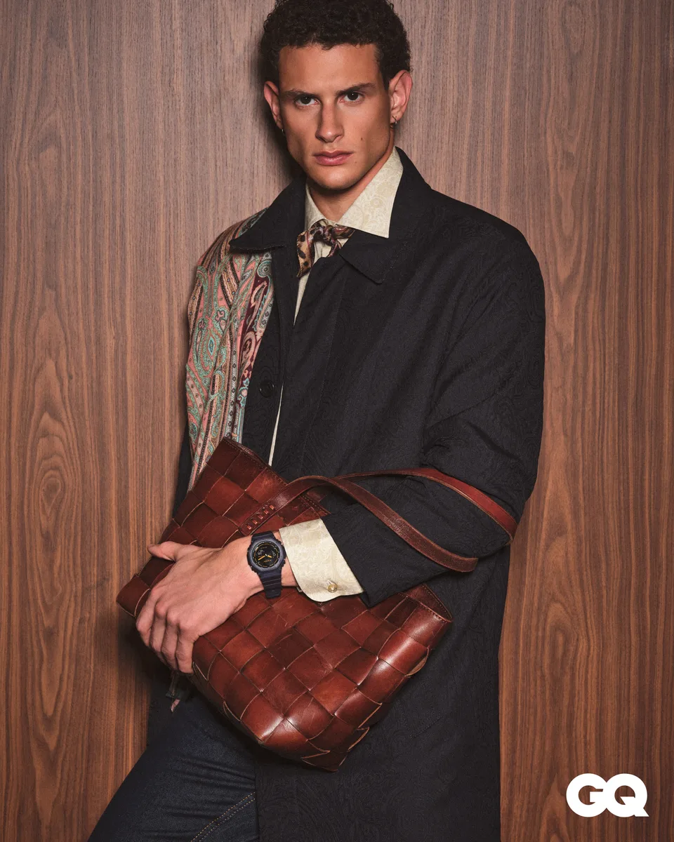 A model with a leather bag wears a watch against a wood-panelled background.