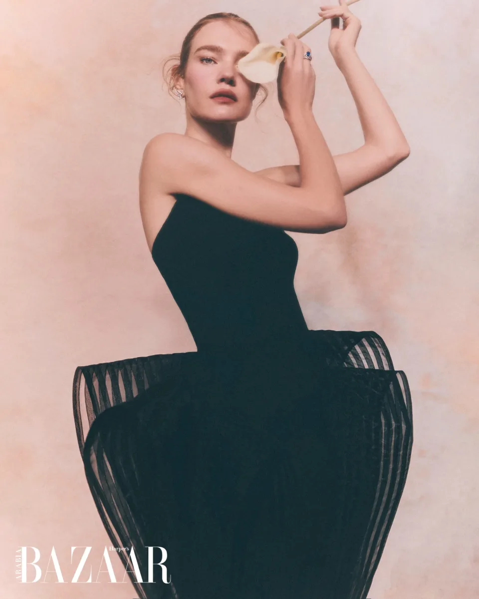 Person in a strapless black dress holding a white flower near the face while seated in a black slatted chair against pink, with Harper's Bazaar text.