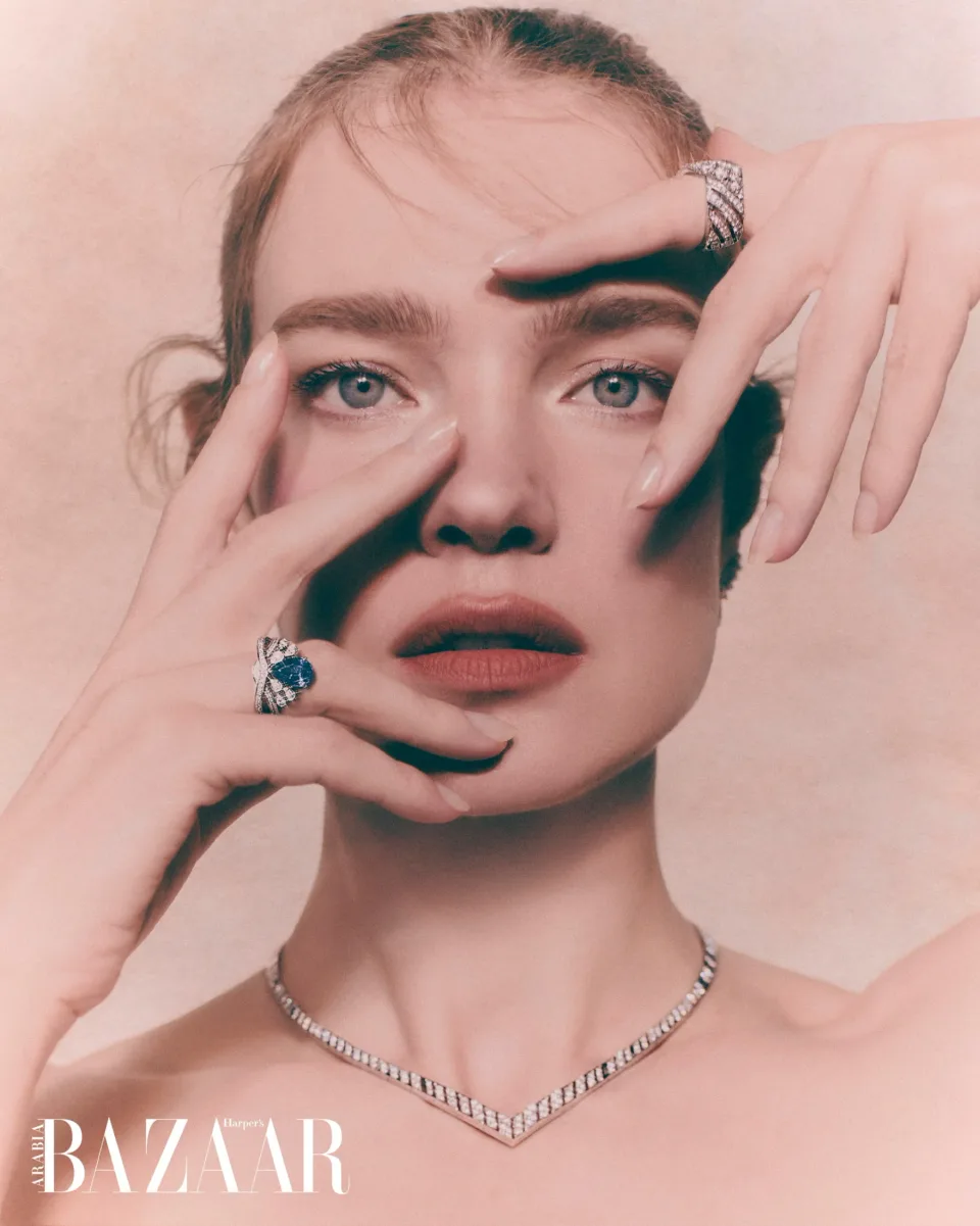 Close-up face framed by hands wearing rings and a necklace against a pink background, with Harper's Bazaar text.