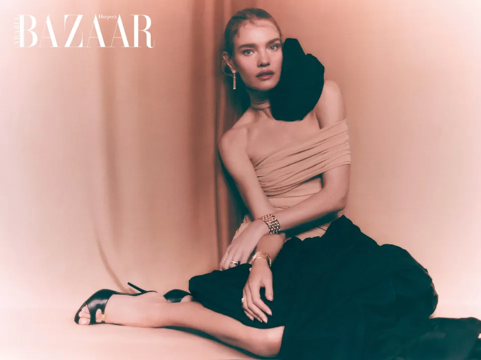 Person seated in a pale pink draped top and black skirt against a pink curtain, wearing earrings and bracelets, with Harper's Bazaar text.