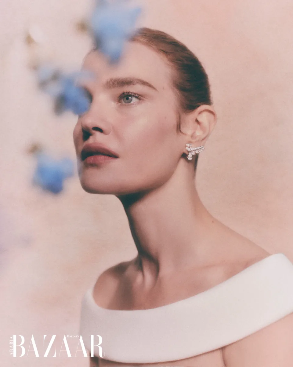 Side-profile close-up with a jeweled earring and white top, partly obscured by blurred blue flowers against pink, with Harper's Bazaar text.