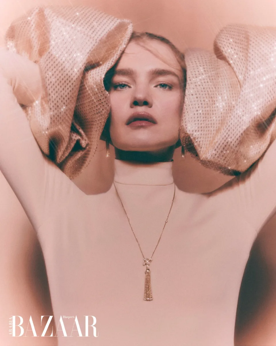 Person in a pale high-neck top with raised arms and shimmering sleeves framing the head, wearing a long pendant against pink.