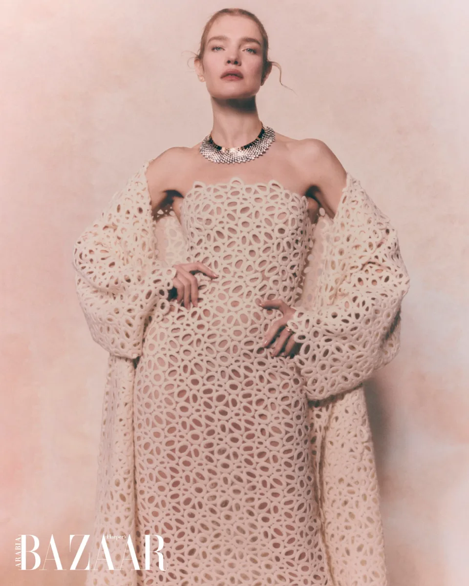 Person in a white textured dress and open patterned coat, wearing a necklace and rings against pink, with Harper's Bazaar text.