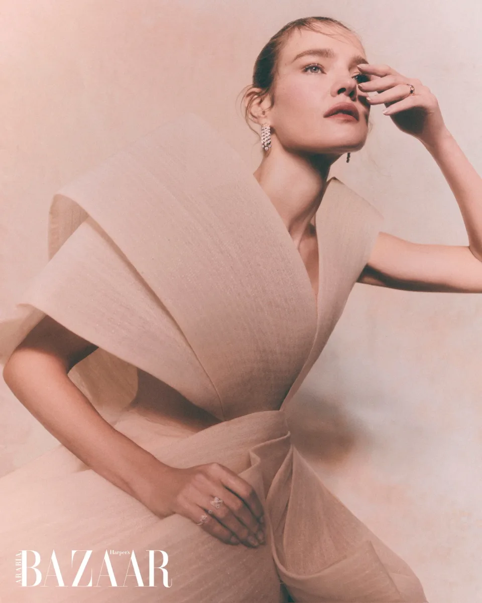 Person in a pale structured draped garment with jeweled earrings and rings, one hand near the face against pink, with Harper's Bazaar text.