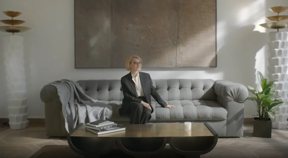 A person sits on a grey sofa in an interior with large artworks, plants and a low coffee table.