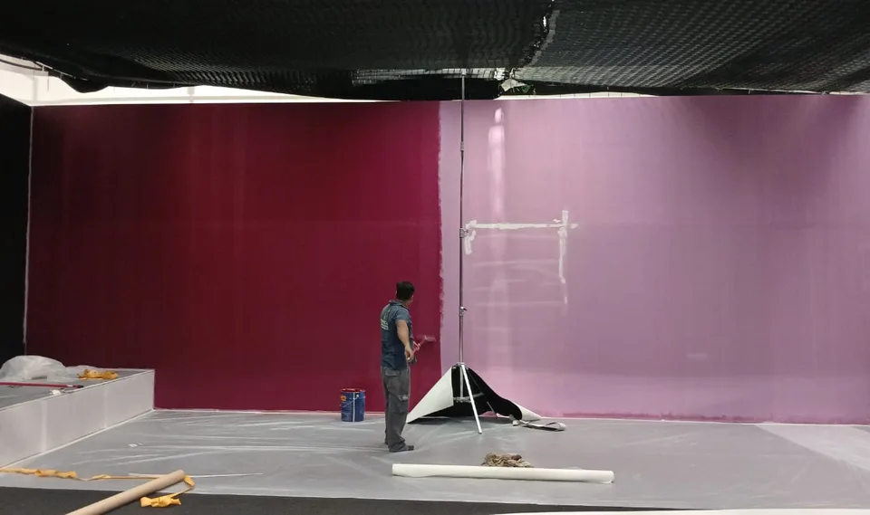 A crew member stands beside the tall set walls as their surfaces are finished in the studio.