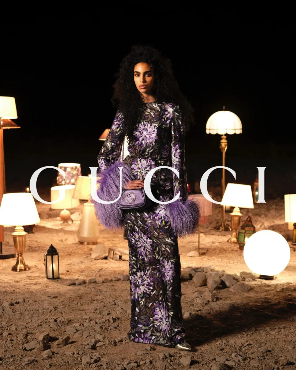 Person in a purple floral outfit with fur-trimmed sleeves and a crossbody handbag standing among lamps on sandy ground, with GUCCI text.