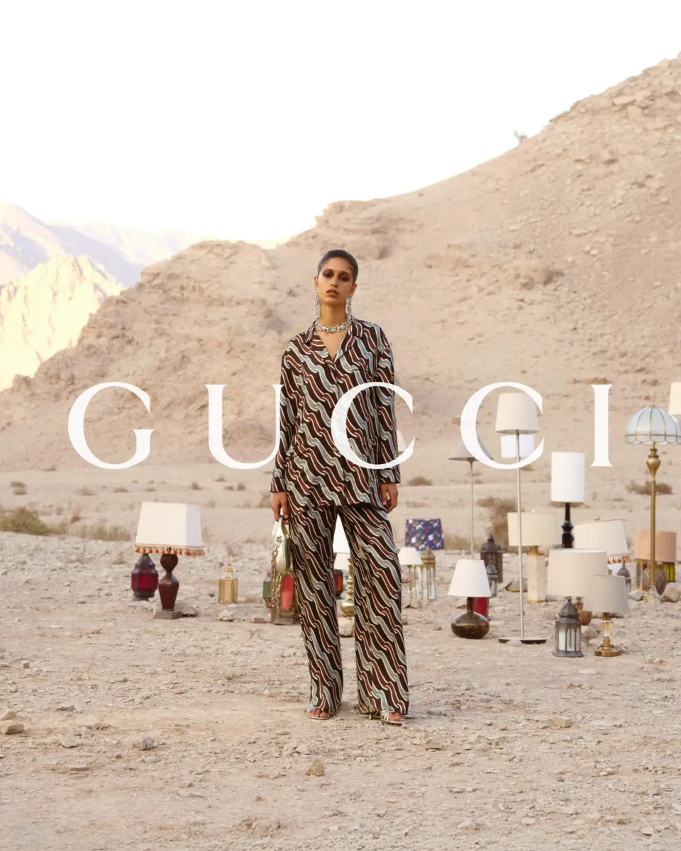 Person in a brown patterned outfit standing among lamps and lanterns on rocky desert ground with mountains behind, with GUCCI text.