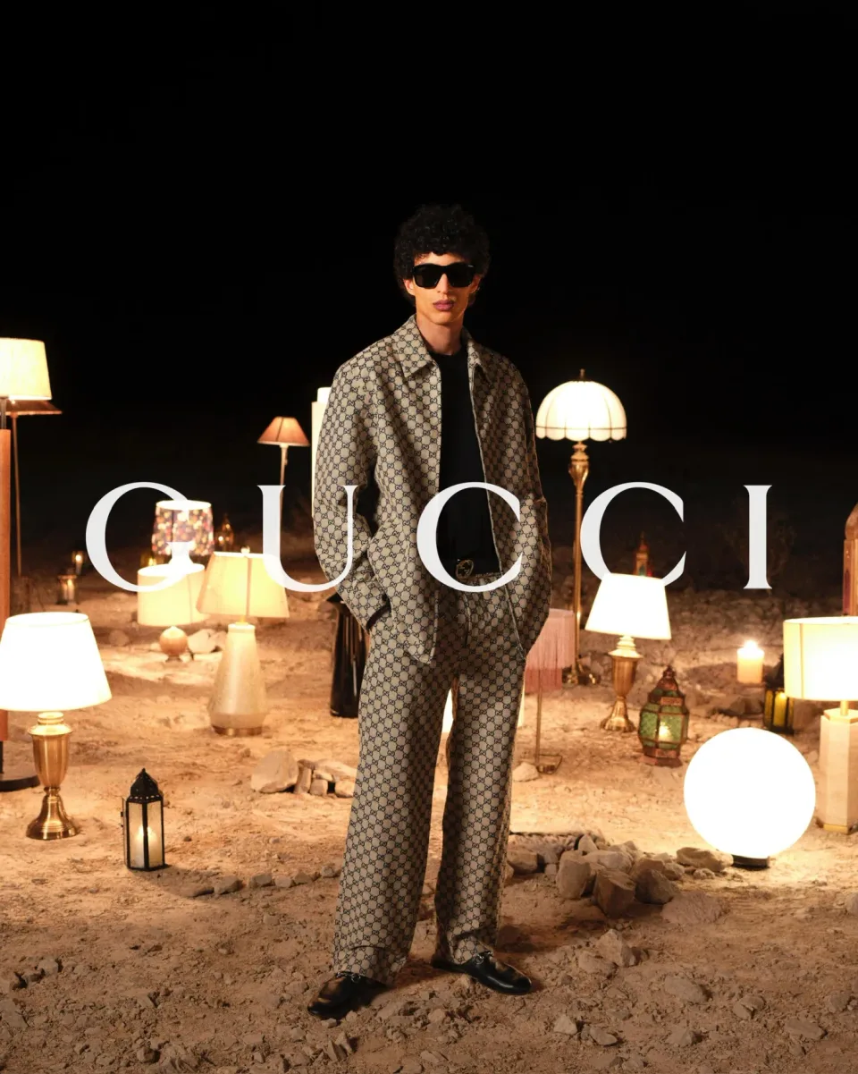 Person in a beige patterned outfit and dark sunglasses standing among lamps on sandy ground at night, with GUCCI text.
