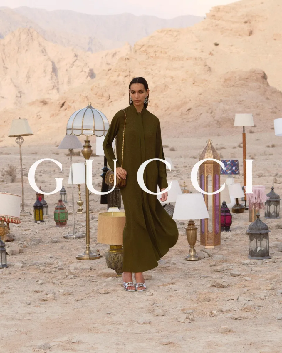 Person in an olive dress carrying a shoulder bag among lamps and lanterns on pale rocky ground, with mountains behind and GUCCI text.