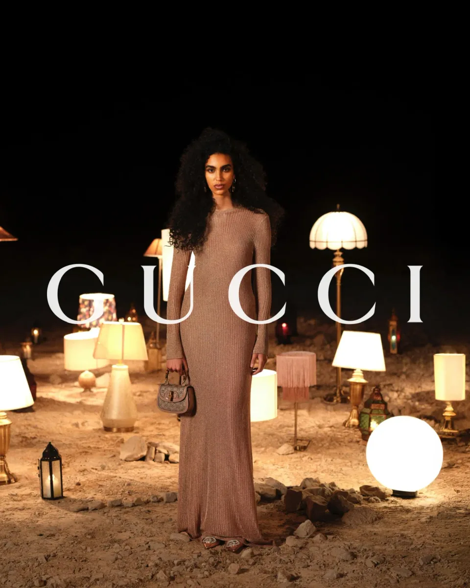 Person in a long ribbed beige dress holding a handbag among lamps on sandy ground at night, with GUCCI text.