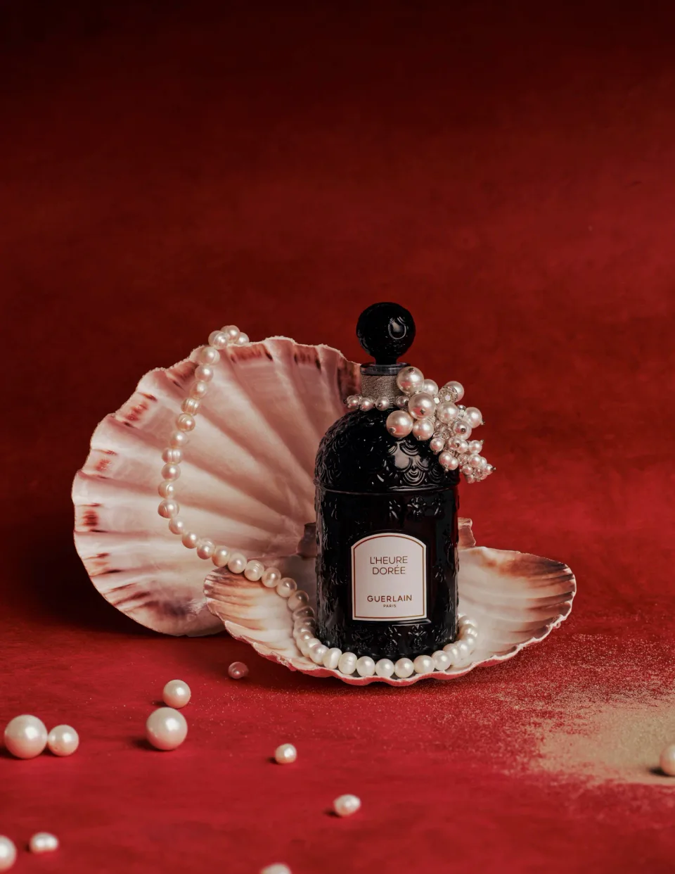 Black Guerlain fragrance bottle on a red set, framed by shells and strands of scattered pearls.