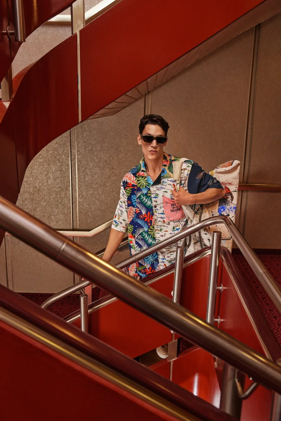 Model in sunglasses and a patterned shirt stands on a red staircase with metal railings.