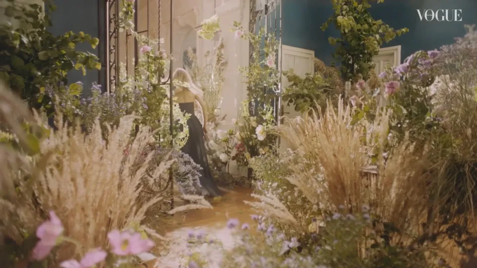 Woman in black stands partly hidden among flowers and tall grasses in a sunlit room.