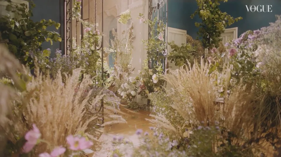 Tall grasses, flowering branches and foliage fill the blue room around a model walking beside the windows.