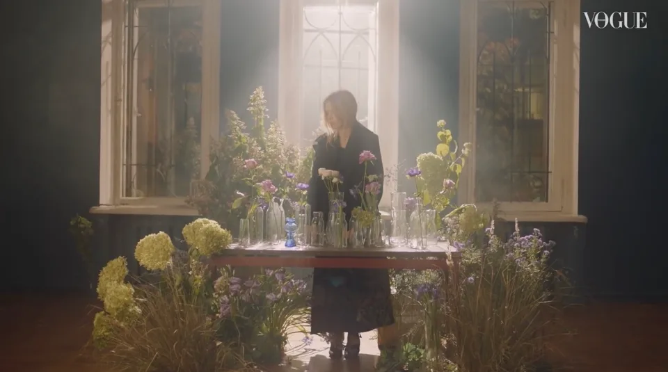 A model stands behind a table of flowers and glass vases, with grasses and blooms growing around its legs.