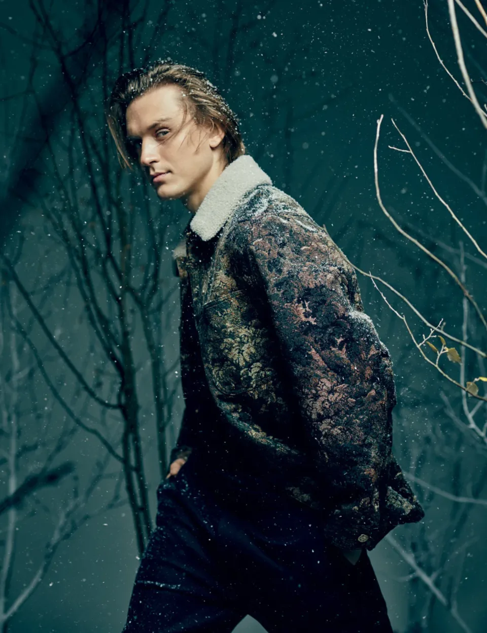 Person in a patterned coat stands among dark winter branches as snow falls.