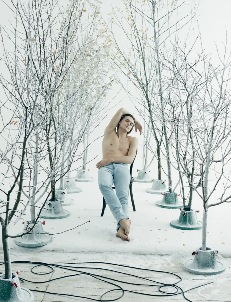 Person seated on a chair between bare trees in a white studio set