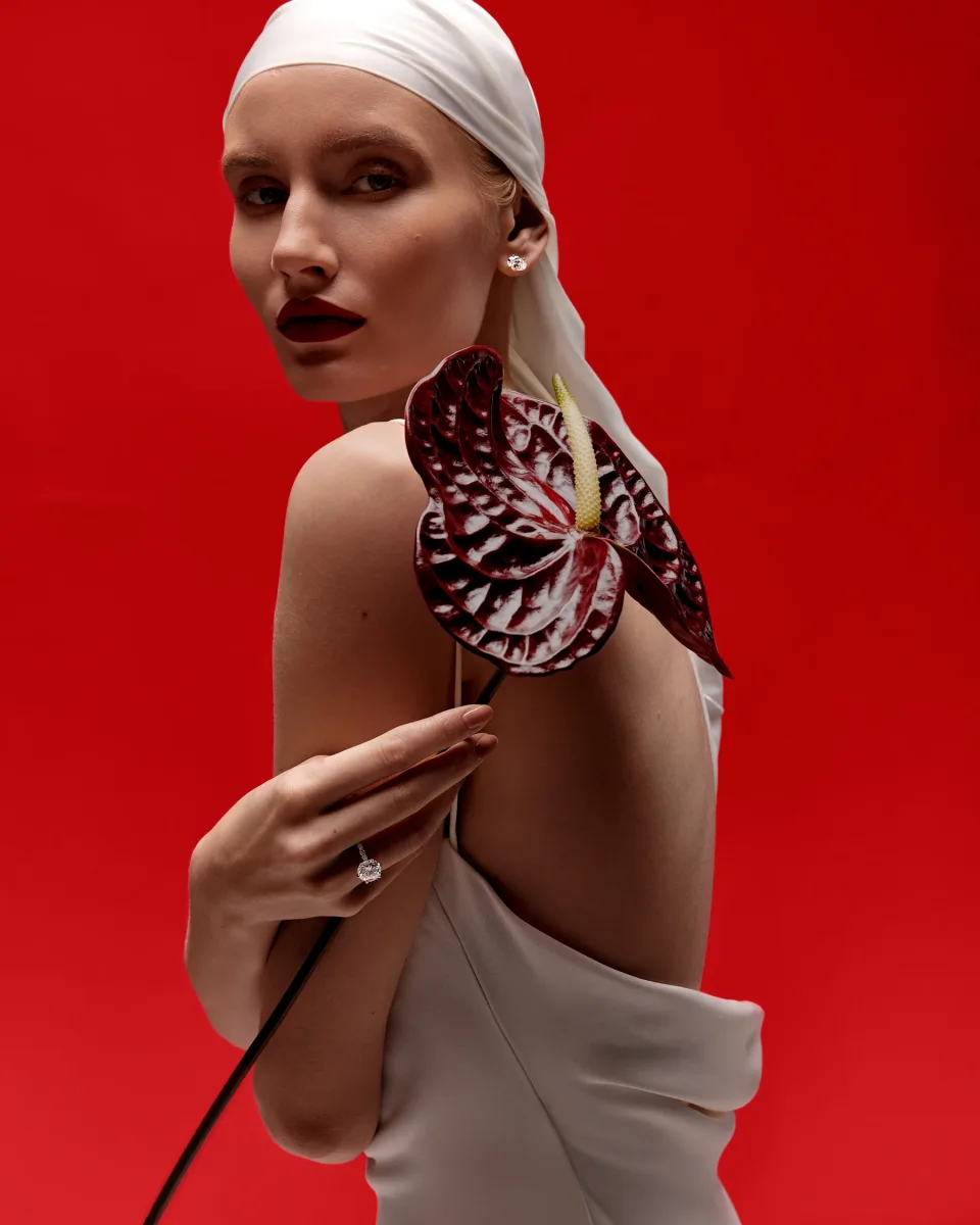 Person in a white head covering holding a large dark red flower against a vivid red background, with rings and an earring visible.
