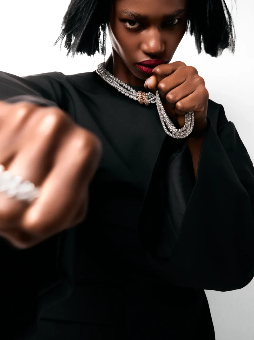 Close view of a person in black holding a jeweled necklace near the collar, with a ringed hand blurred in the foreground against white.