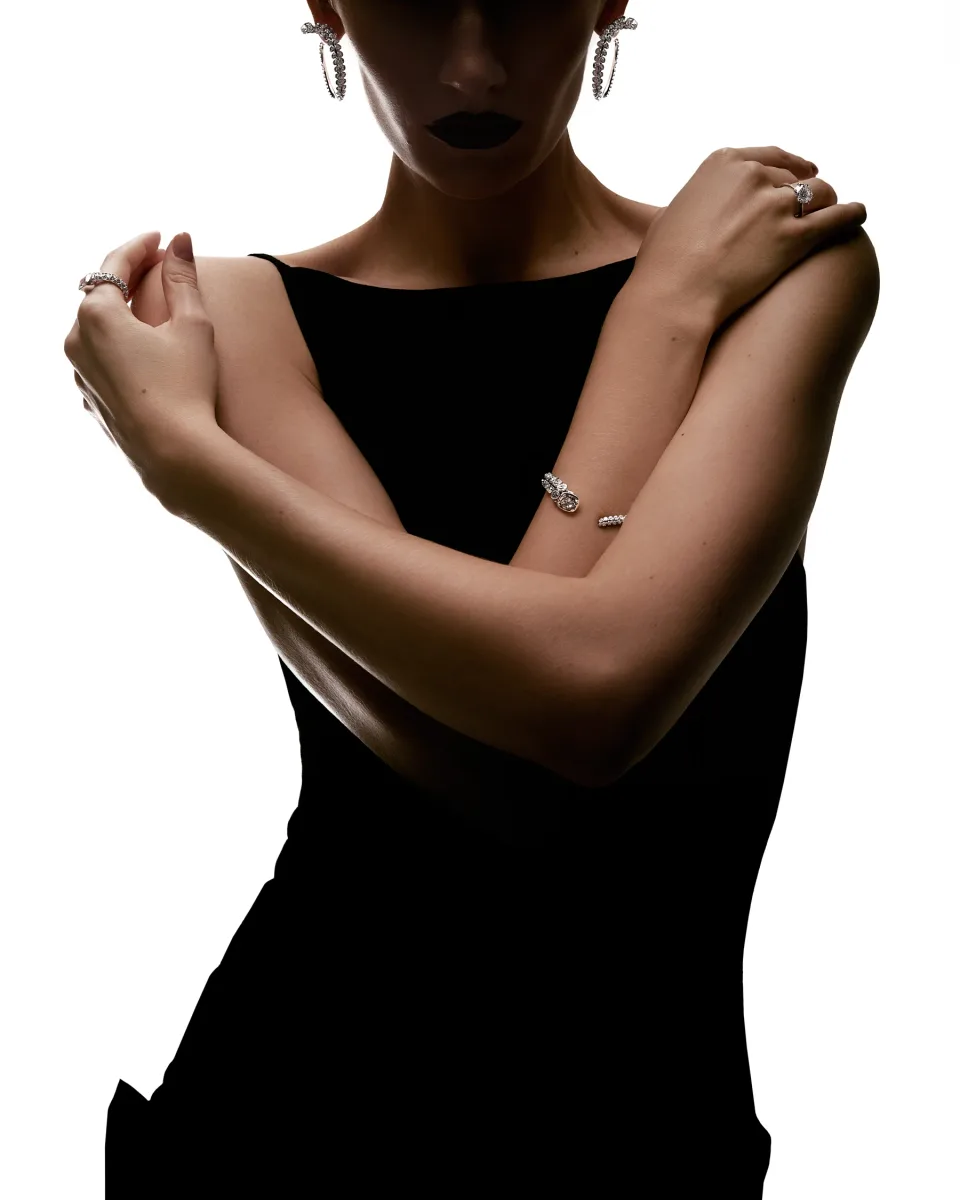 Cropped person in a black sleeveless top with crossed arms, jeweled rings, bracelet and earrings against a white background.