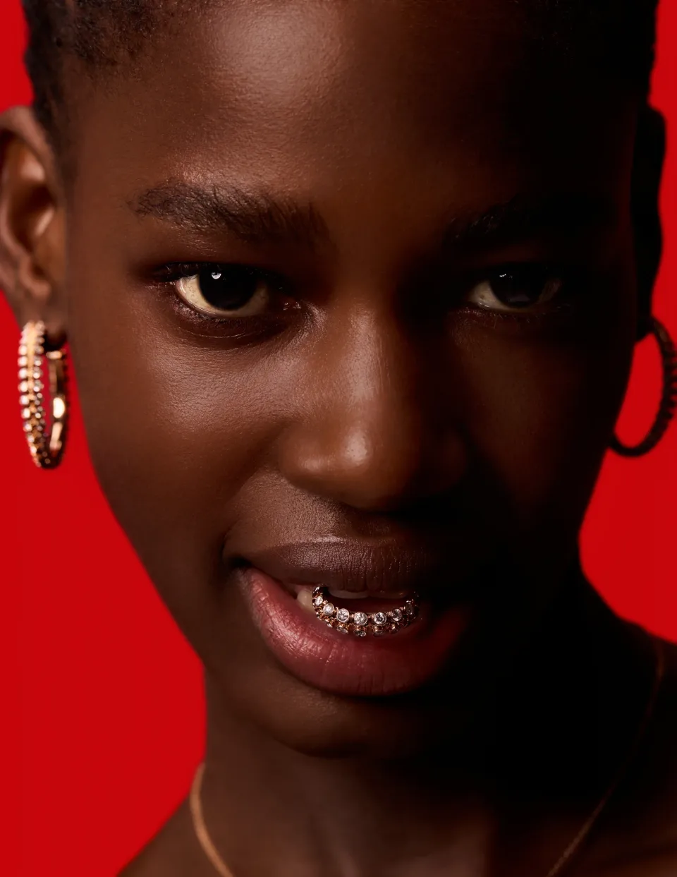 Close-up face against a red background with hoop earrings and a jeweled ornament at the lower teeth.