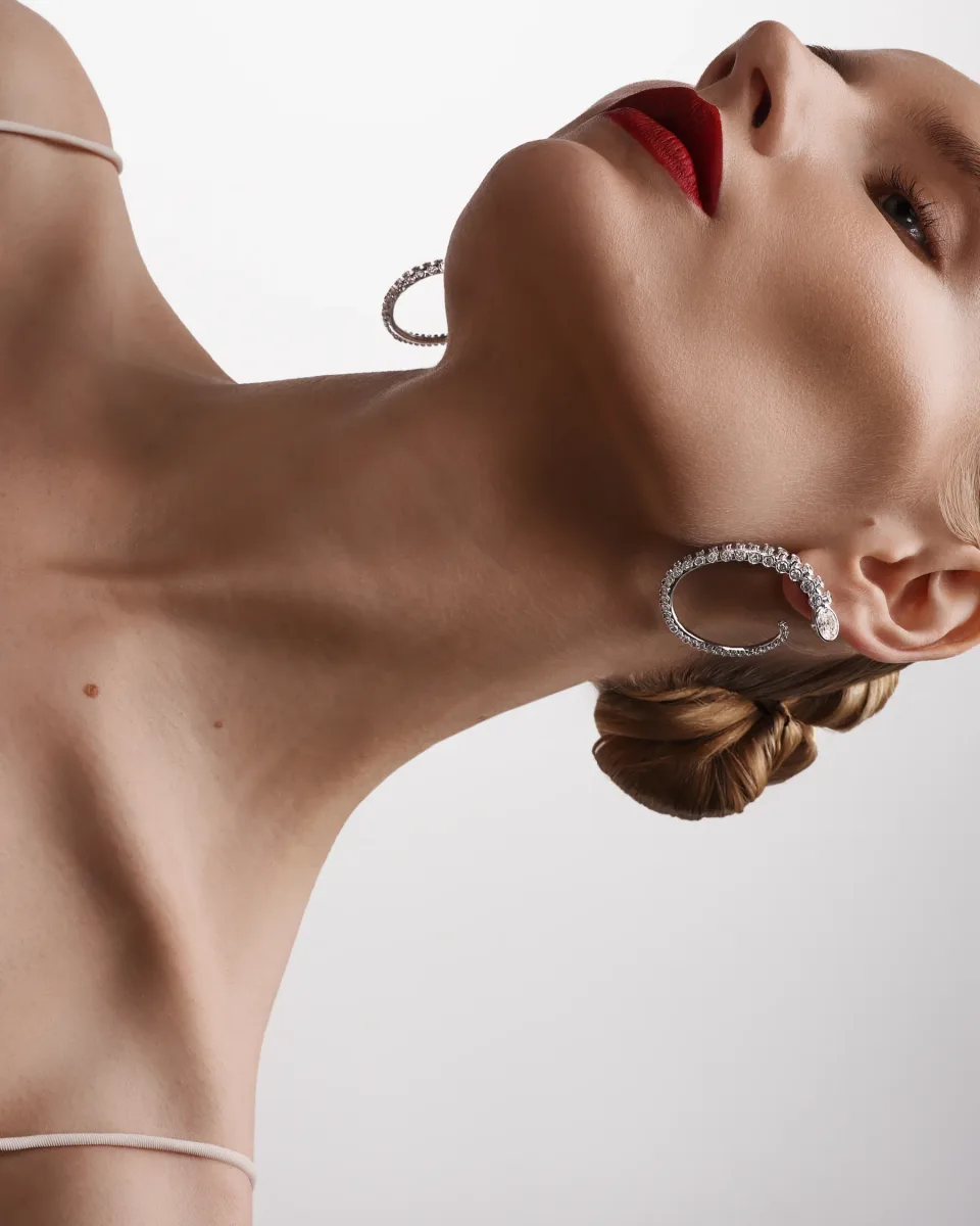 Angled close-up of a person with red lipstick and jeweled hoop earrings against white, with a white strap visible.