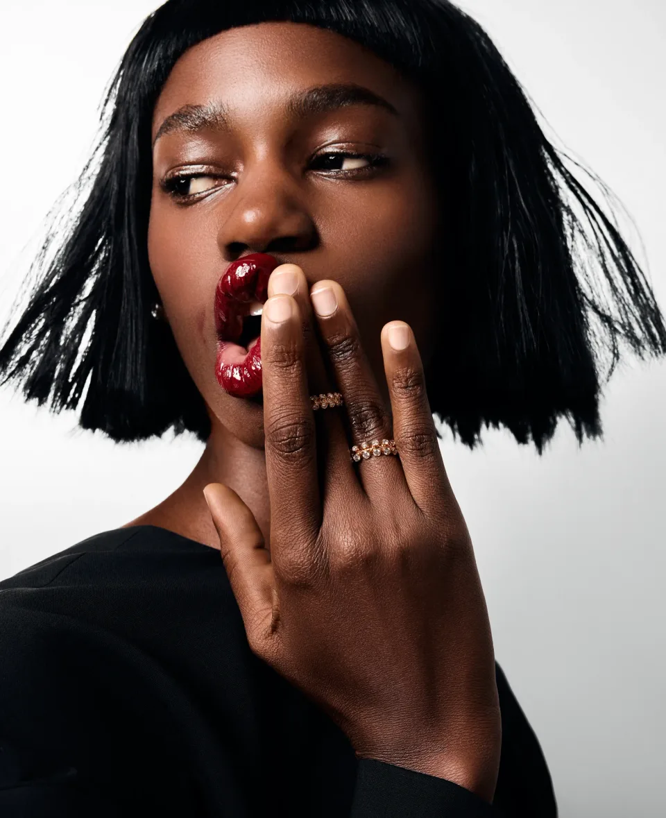 Close-up of a person with a dark bob haircut touching glossy dark red lips, wearing rings against a white background.