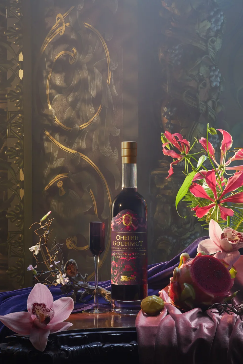 Onegin Gourmet bottle surrounded by pink flowers, fruit and layered purple fabric against a dark ornamental backdrop.