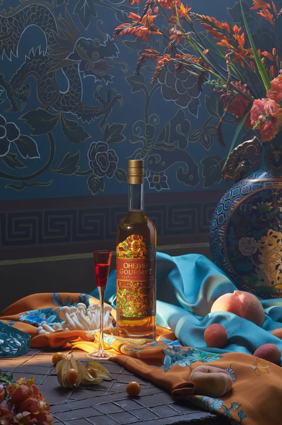 Onegin Gourmet bottle and red stemmed glass on a table with blue and orange fabrics, fruit and flowers against a patterned blue wall.