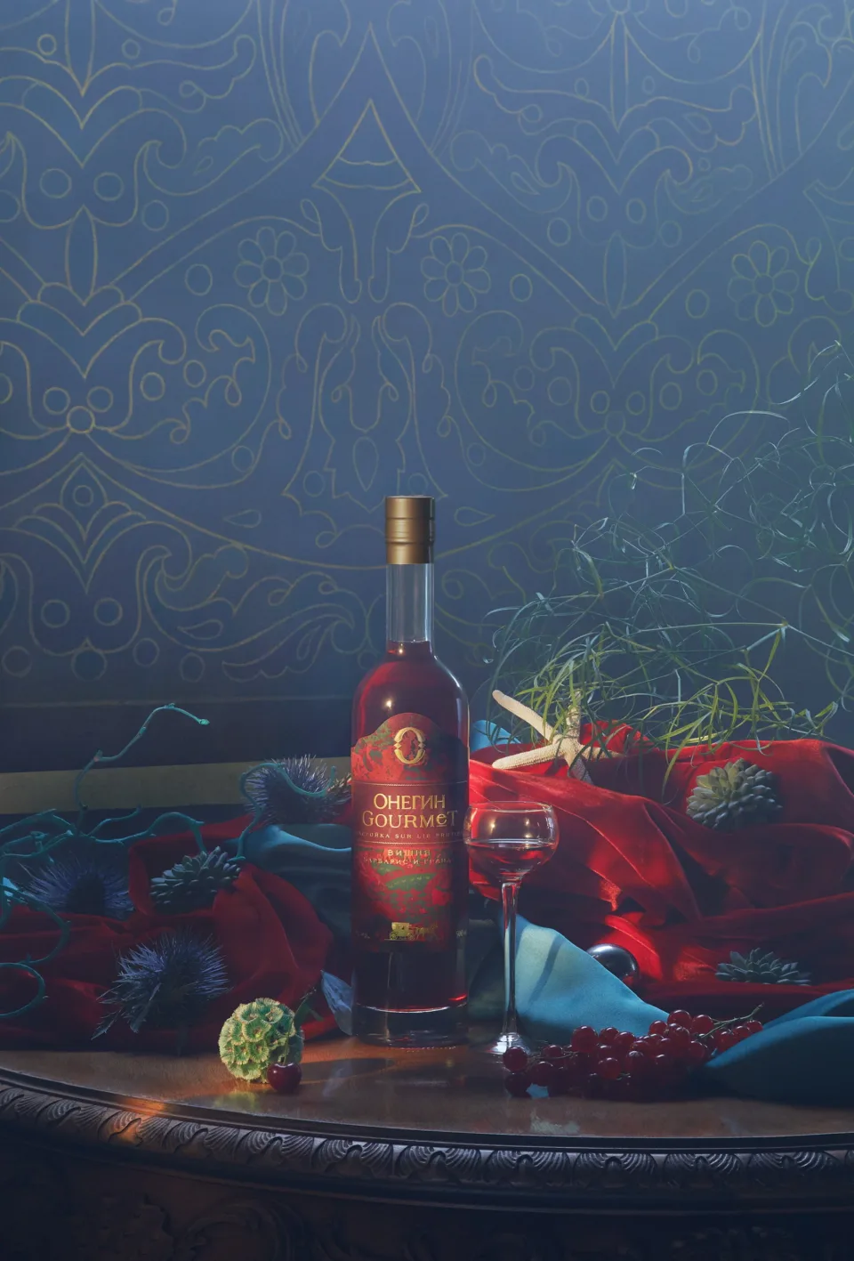 Onegin Gourmet bottle and stemmed glass among red and blue draped fabrics, fruit and flowers against a blue patterned wall.