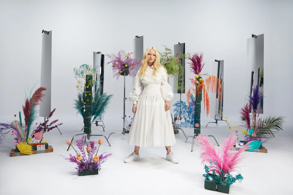 A model stands among colourful flowers, plinths and freestanding studio lights.