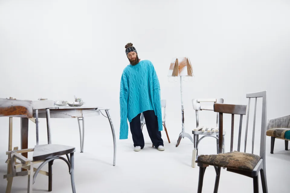 A model stands among chairs, a table and a floor lamp in a white studio.