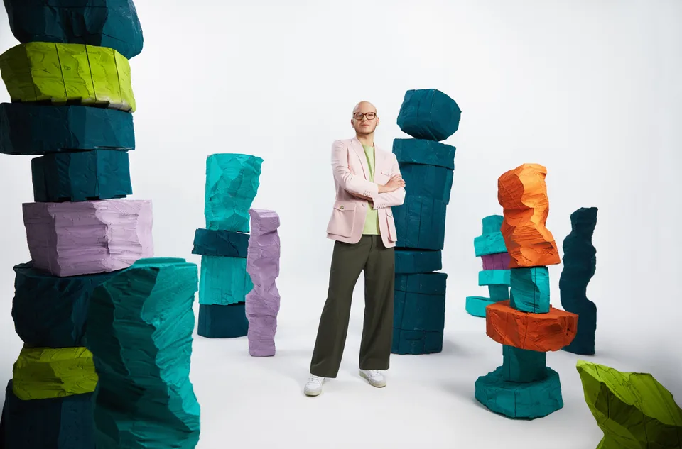 A model stands among tall stacks of textured sculptural forms in green, orange, pink and blue.