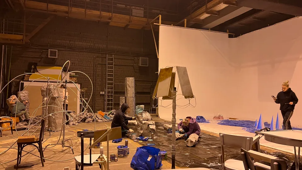 Backstage view shows several scenic settings and props being prepared across a large studio.