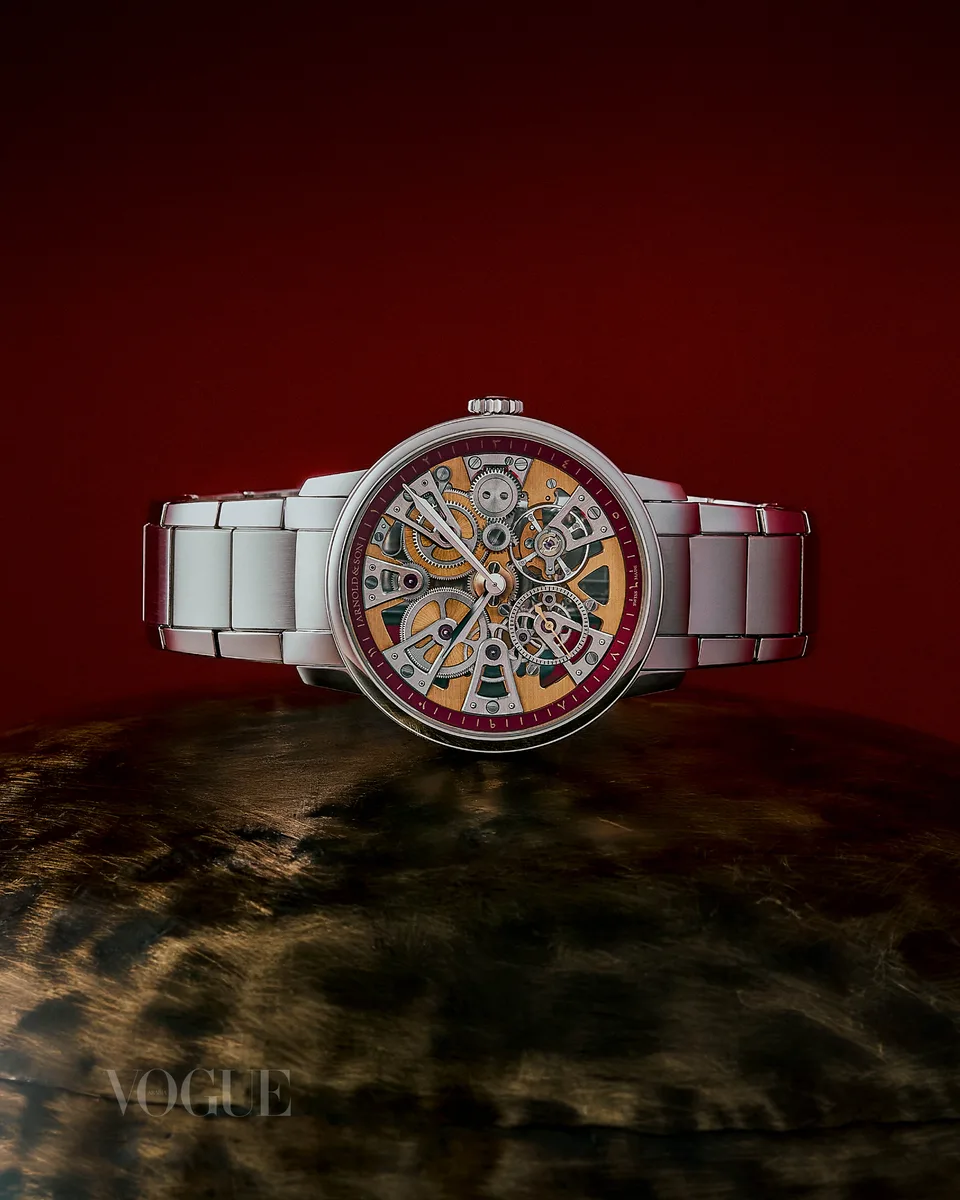 A skeleton-dial watch sits above a textured surface against a red backdrop.
