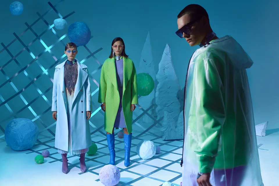 A model in a lime green jacket stands in front of the blue set and grid shadows.