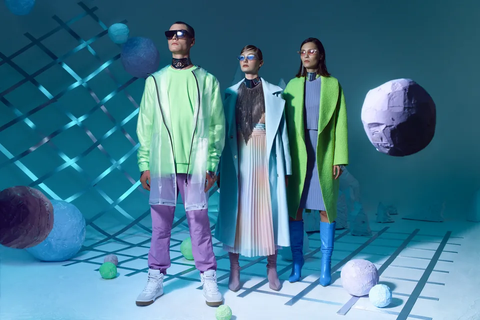 Three models stand in a blue sculptural set with coloured spheres and a tilted grid.
